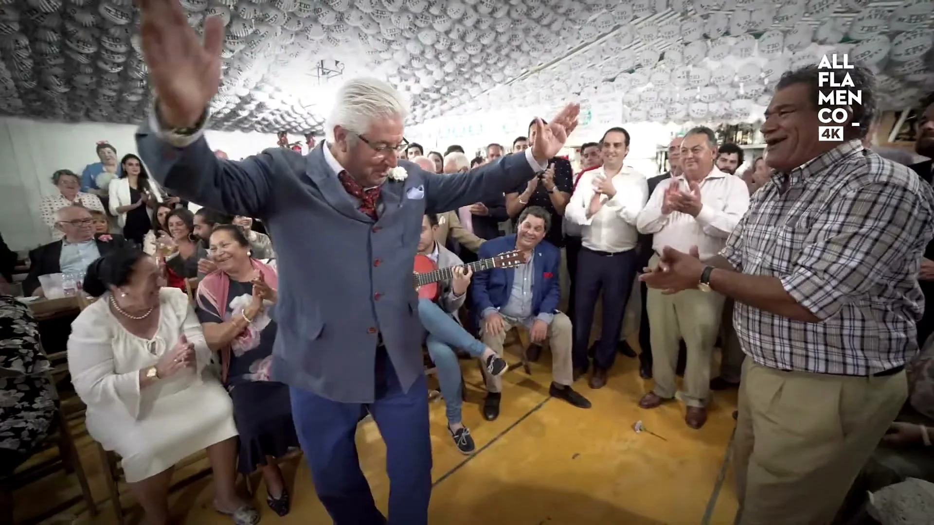 A man dancing sevillanas in the middle of a feria marquee, arms open, with the circle of seated people clapping around him