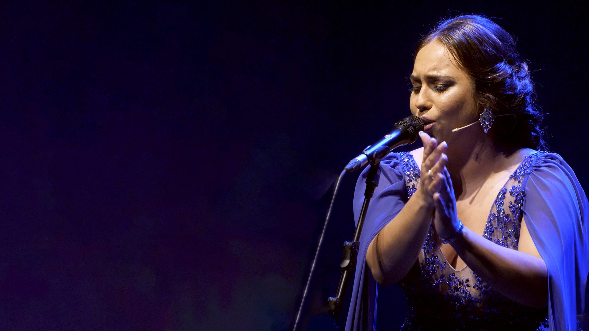María Terremoto con vestido azul, cantando ante el micrófono con las manos juntas