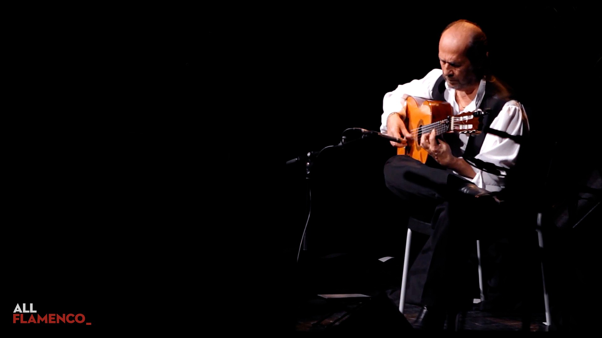 Paco de Lucía sentado y solo en un escenario oscuro, tocando la guitarra