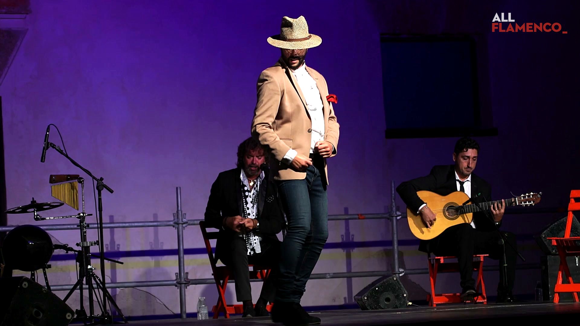 David Nieto, in a straw hat and a light jacket, dances por guajira on a stage lit in blue
