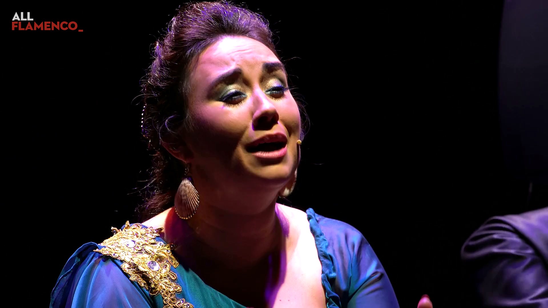 Close-up of Encarna Anillo singing, wearing a shell earring and a blue dress, against a black background