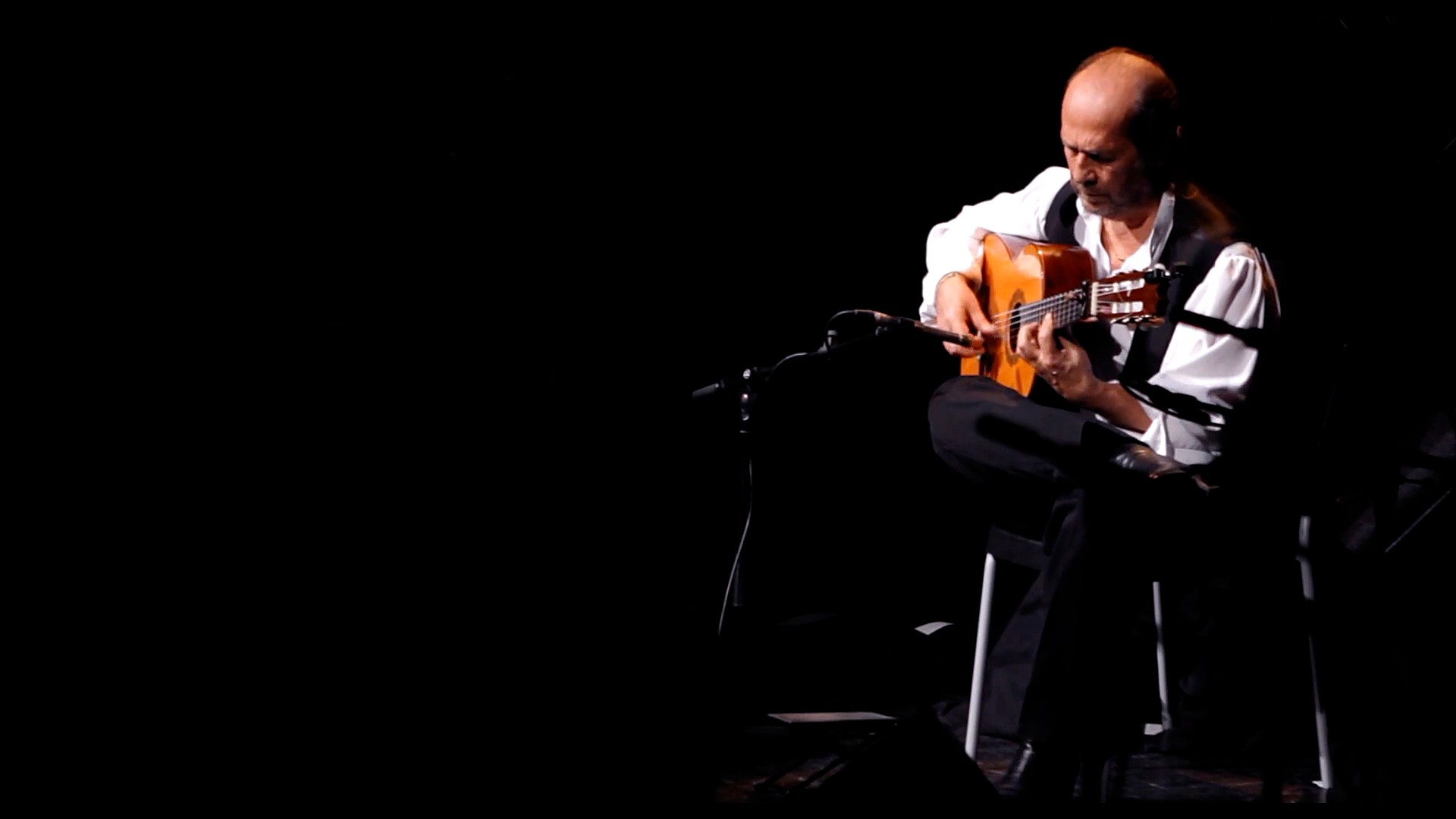 Paco de Lucía sentado y solo en un escenario oscuro, tocando la guitarra