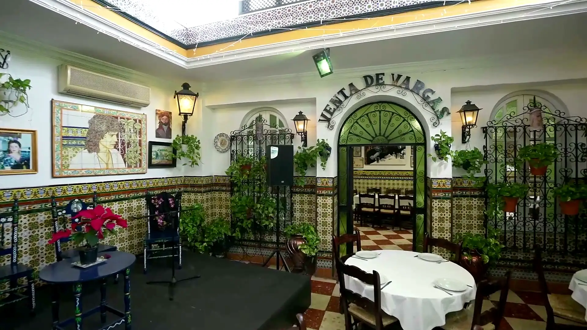 The courtyard of Venta de Vargas in San Fernando, with the wrought-iron «Venta de Vargas» sign above the green arched doorway, tiled dado, hanging flowerpots, chequered floor and a table laid with a white cloth